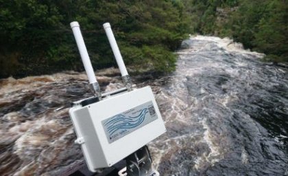A white camera-looking object with two antennas sits above fast flowing brown water with bushland in the background.
